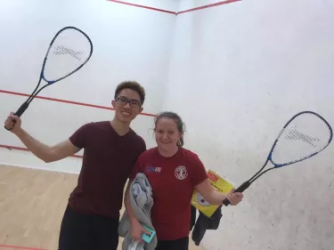 Ellie with a friend, both holding up their racquets and smiling, in the Christ's College squash court